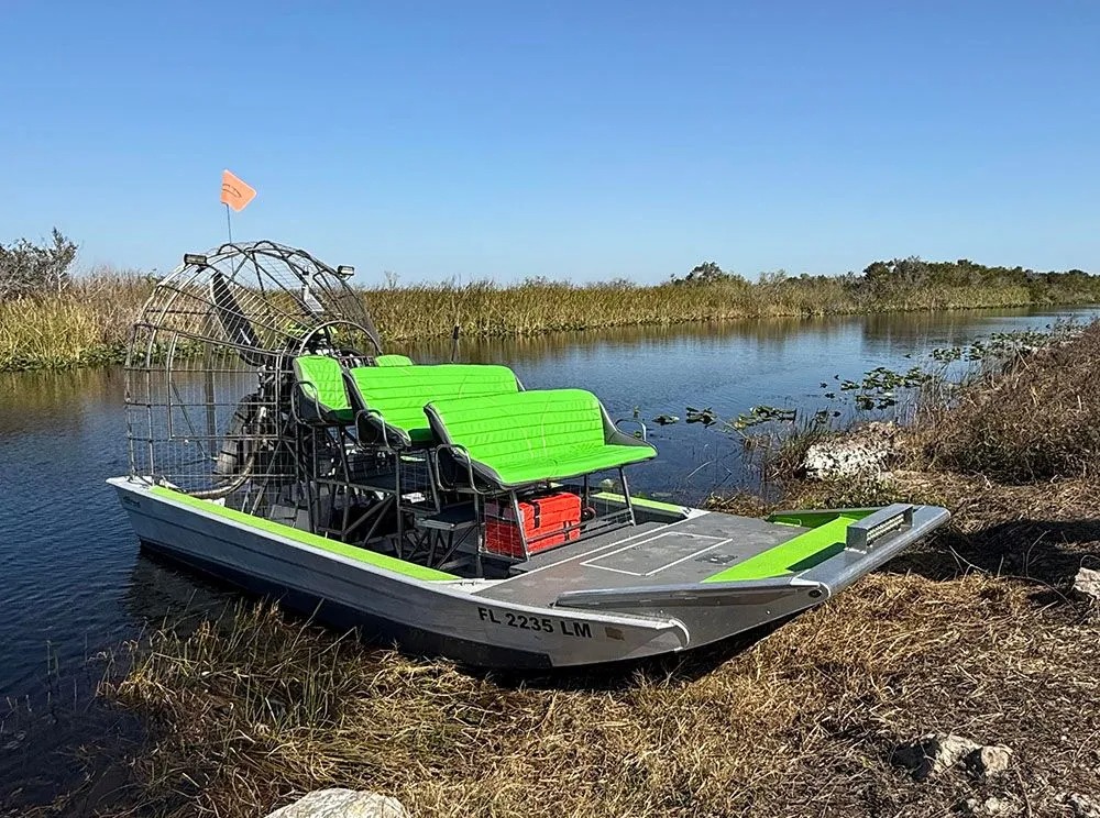 Airboat with green seats docked by a grassy waterway under a clear blue sky.